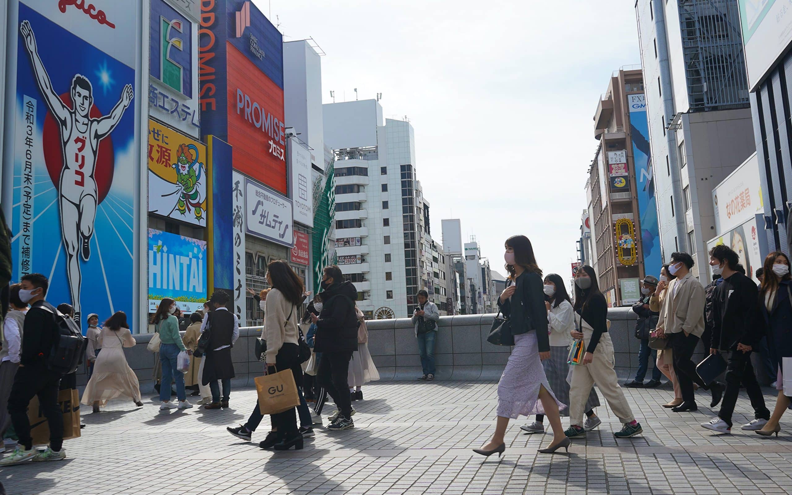 Photo of Osaka, Japan - April 1, 2021: a crowd of people wearing masks passing Ebisu Bridge in Osaka with the famous Glico sign and other billboards in the background.