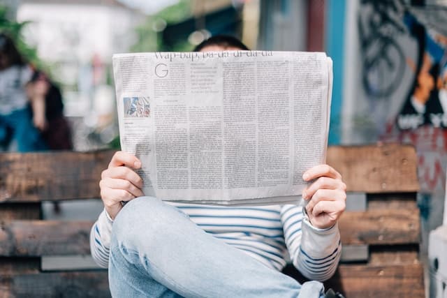 A woman holding a newspaper up and covering her face while reading on a bench. Photo by Roman Kraft on Unsplash