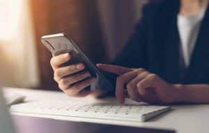 Woman using smartphone - her face is not visible. The focus is on her hands, arm, phone and a keyboard on a desk