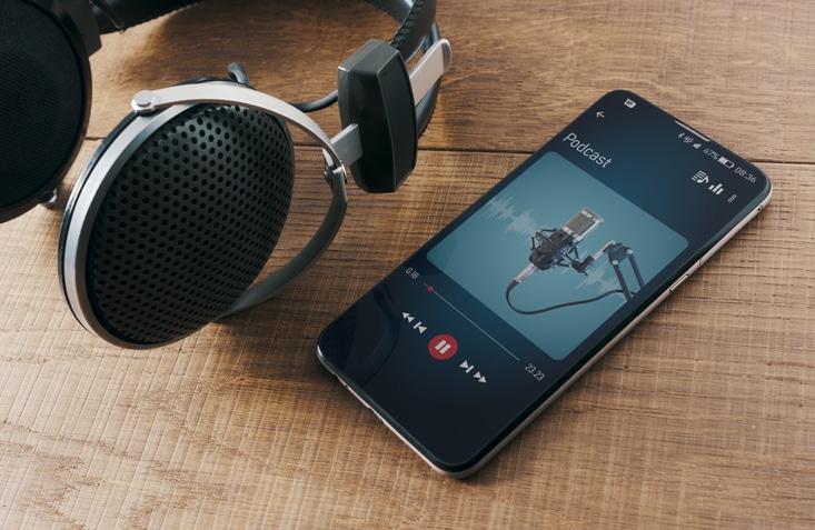 Photo of A pair of headphones and a smartphone on a wooden table. On the display you see the player, and a Podcast playing.