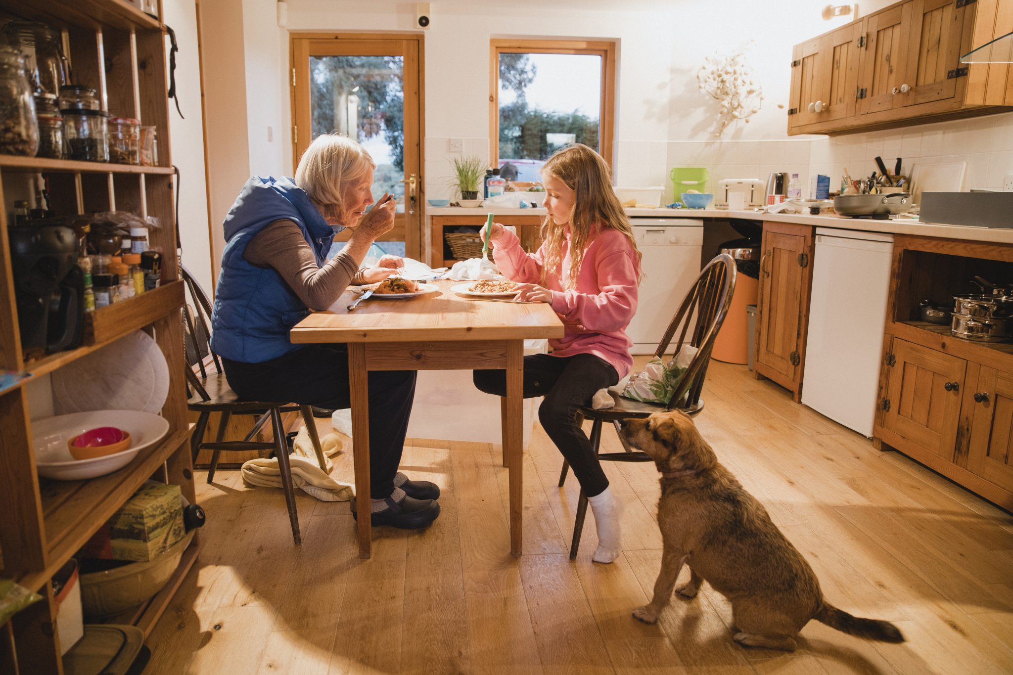 Side view of a grandmother and her granddaughter sitting at the dining table. They are both eating their meal. On the floor next to the little girl there is a pet dog sitting waiting for some food.